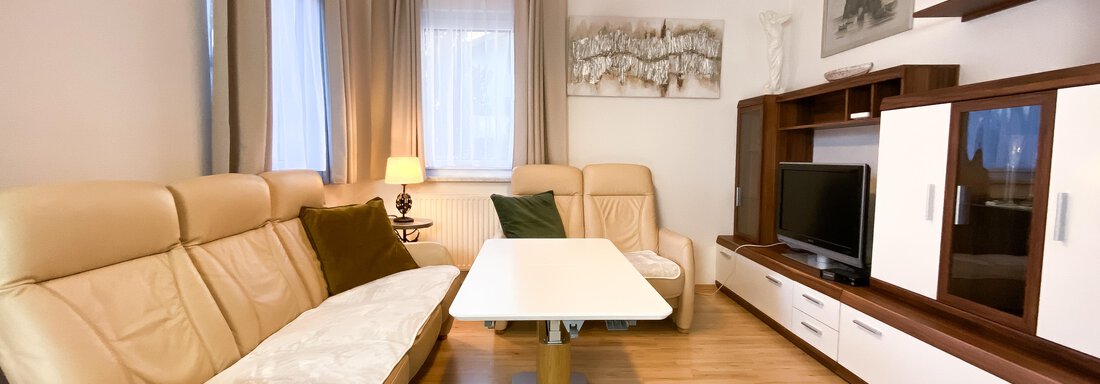 Ein modernes Wohnzimmer mit einer gemütlichen beigen Couch und einem stilvollen Tisch. Der Raum ist hell und freundlich, mit Holzfußboden und großen Fenstern. - Ferienhaus Neurauter - Haiming