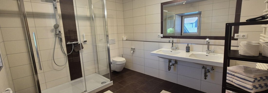 A modern bathroom with a walk-in shower and two sinks. The walls are tiled in light colors and the floor is fitted with dark tiles. - Landhaus Waldesruh - Haiming