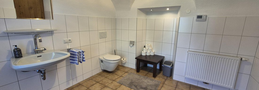 A modern bathroom with white tiles and a large, bright area. There is a toilet, a sink, and a small table. - Landhaus Waldesruh - Haiming