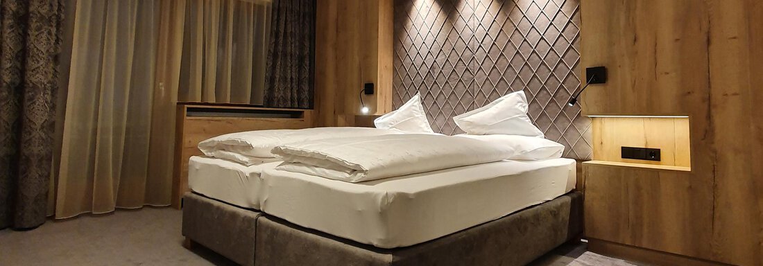 A modern bedroom with a double bed and soft lighting. The walls are designed in warm wood tones, and large windows provide light. - Alpenhotel Laurin - Hochgurgl