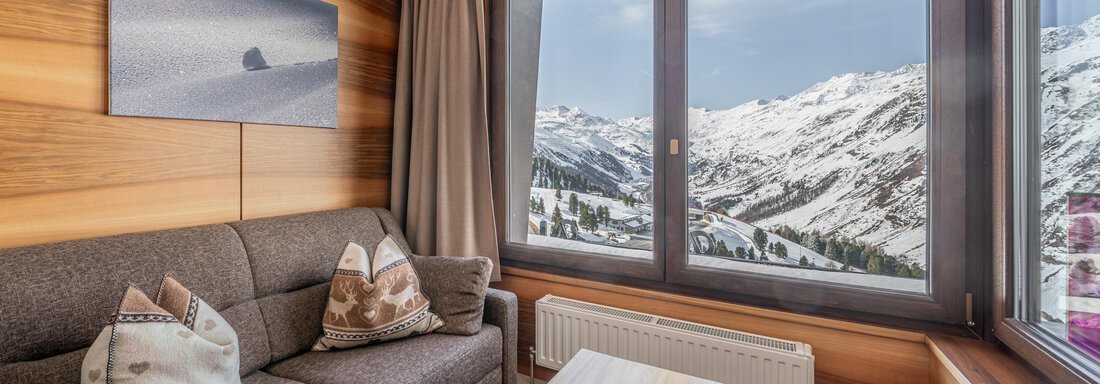 A cozy living room with a gray couch and a table. Through large windows, the snowy mountain landscape is visible. - Hotel Angerer Alm - Hochgurgl