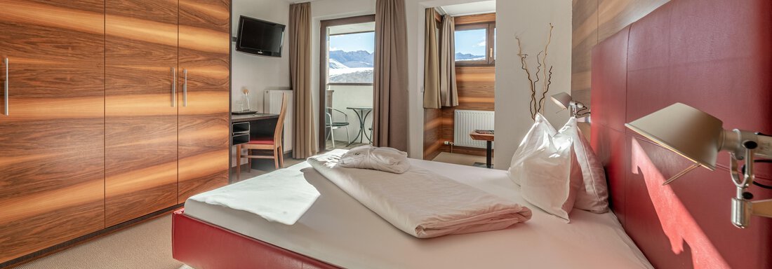 web-angerer-alm-zimmer103--2 A modern bedroom with a red bed and a wood-colored wardrobe. Windows let in plenty of daylight and offer a view outside. - Hotel Angerer Alm - Hochgurgl