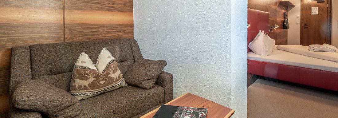 A cozy room with a sofa and a small wooden table. In the background, you can see a bed and a decorative wall with a picture. - Hotel Angerer Alm - Hochgurgl