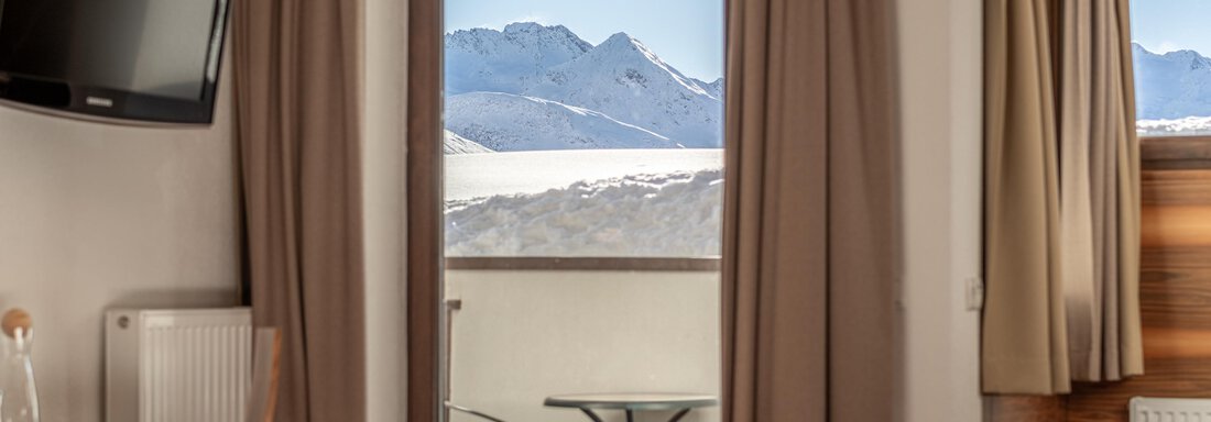A cozy room with a view of snow-covered mountains. The window is open and shows a balcony with chairs. - Hotel Angerer Alm - Hochgurgl