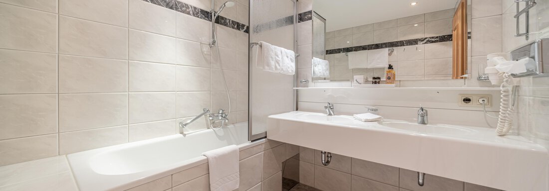 A modern bathroom with a bathtub and a large sink. The walls are brightly tiled and there is plenty of light from the ceiling lights. - Hotel Angerer Alm - Hochgurgl