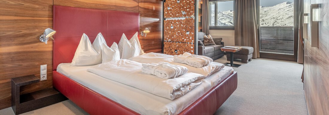 web-angerer-alm-zimmer-221- A modern bedroom with a red bed and elegant wood paneling. The room has large windows that let in plenty of light and offer a view outside. - Hotel Angerer Alm - Hochgurgl