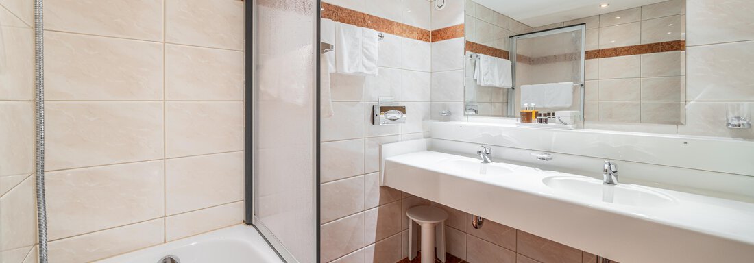 A modern bathroom with a bathtub and a shower. The room is bright and features tiled walls as well as a large mirror. - Hotel Angerer Alm - Hochgurgl