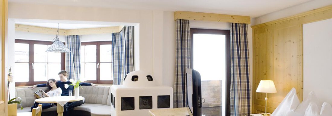 A cozy room with wooden decor and large windows. There is a dining area, a seating corner, and a double bed. - Hotel Angerer Alm - Hochgurgl