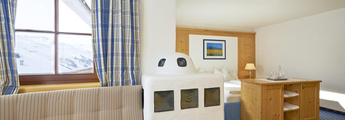 A cozy room with a seating area and a traditional stove. Large windows provide plenty of daylight and a view of the mountains. - Hotel Angerer Alm - Hochgurgl