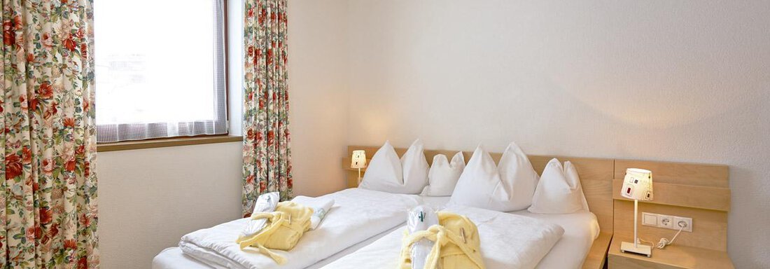 A cozy bedroom with two beds and fresh white bed sheets. The windows are decorated with floral curtains. - Hotel Angerer Alm - Hochgurgl