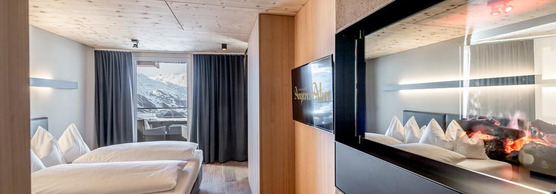 A modern hotel room with a comfortable bed and a cozy fireplace. Large windows offer a view of the mountains and let in plenty of light. - Hotel Angerer Alm - Hochgurgl