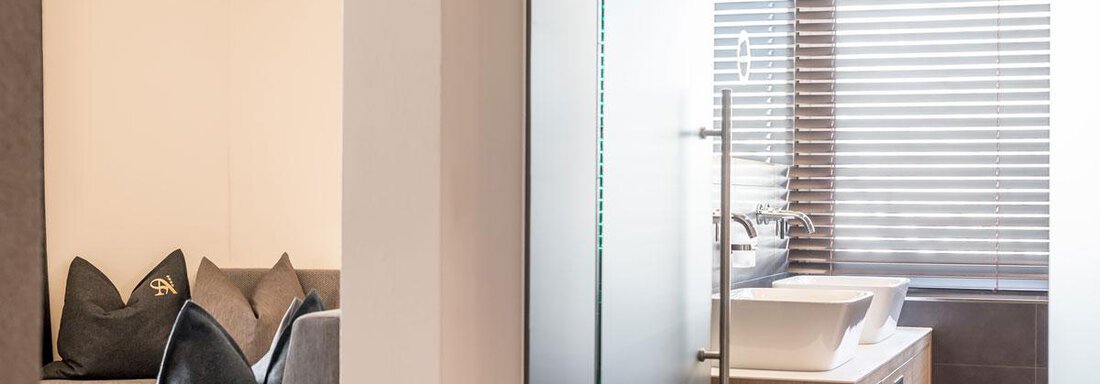 A modern bathroom with a sink and a glass shower. In the background, there is a cozy living area with comfortable cushions. - Hotel Angerer Alm - Hochgurgl