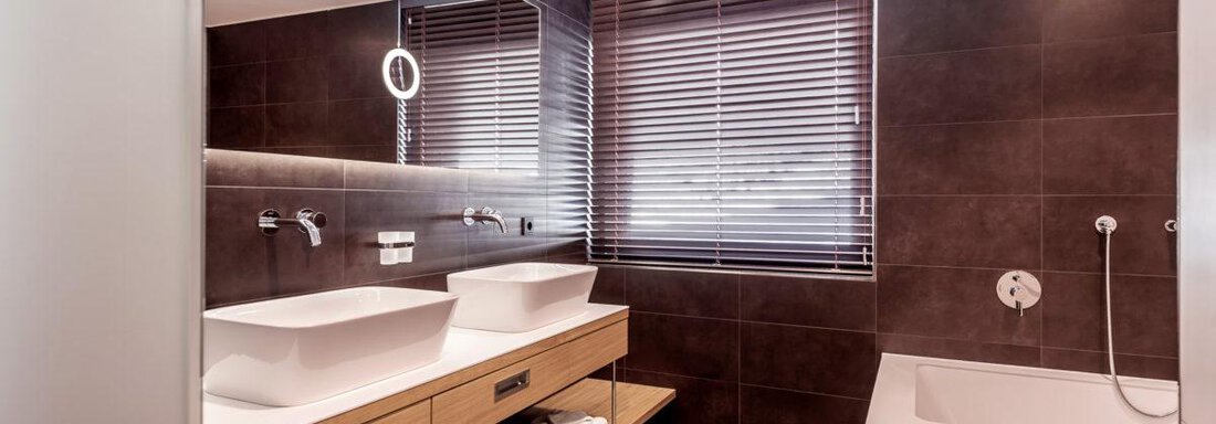 A modern bathroom with wooden cabinets and two sinks. The walls are in dark colors and there is plenty of daylight coming through the windows. - Hotel Angerer Alm - Hochgurgl