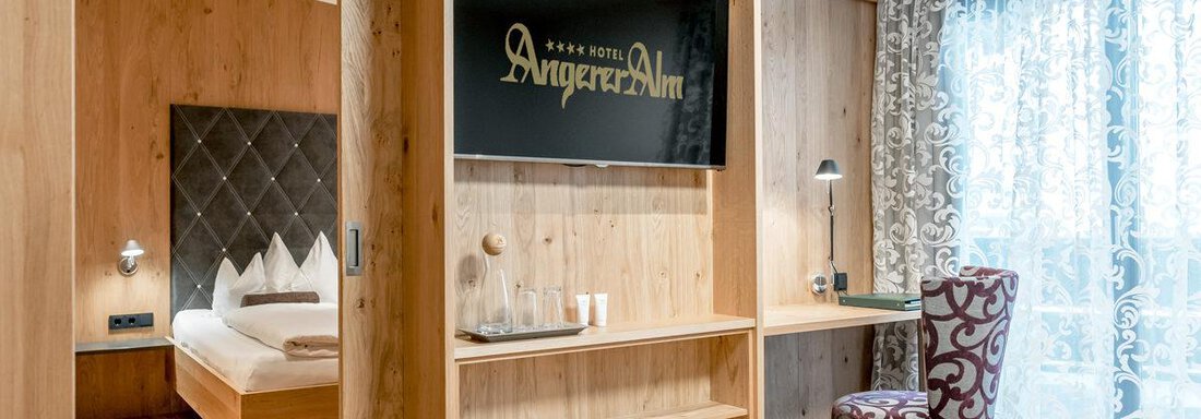 Hotel-Angerer-Alm_Juniorsuite_Teilansicht-1198x799 Modern hotel room with wooden walls and a comfortable bed. A cozy workspace and plenty of daylight through the window create an inviting atmosphere. - Hotel Angerer Alm - Hochgurgl