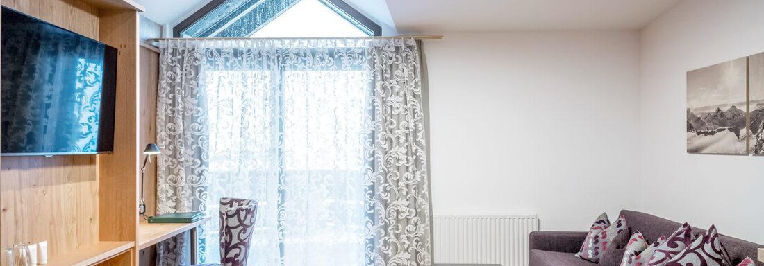 A cozy living room with a modern couch and a table.  
Large windows with curtains let in a lot of light. - Hotel Angerer Alm - Hochgurgl
