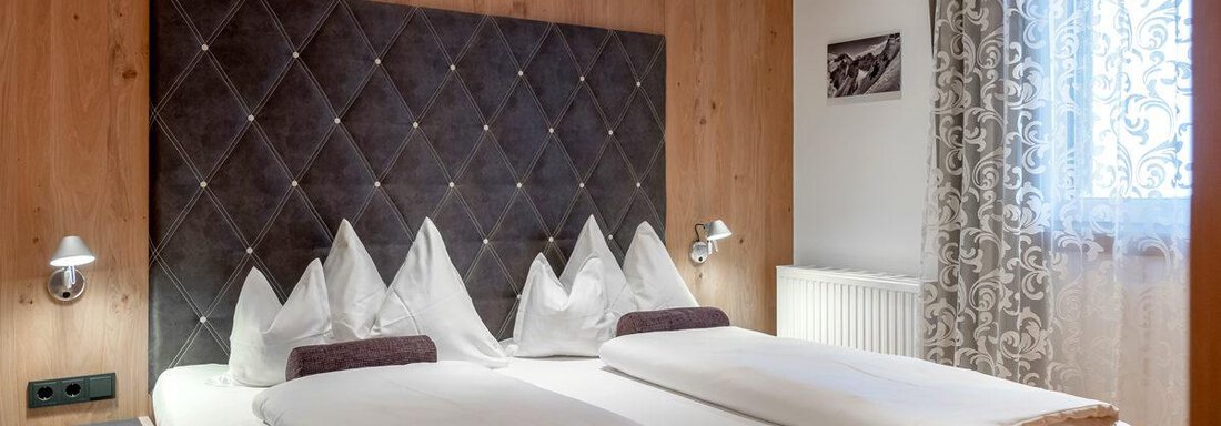 A cozy bedroom with a large bed and elegant bedding. The walls are made of warm wood, and the window lets in plenty of daylight. - Hotel Angerer Alm - Hochgurgl
