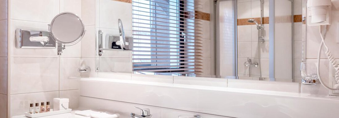 A modern bathroom with two sinks and a large mirror. The shower is separated by a glass wall, and light comes into the room through blinds. - Hotel Angerer Alm - Hochgurgl