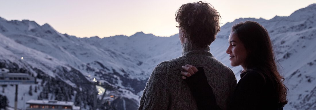 Ein Paar steht auf einem Balkon und blickt auf eine verschneite Berglandschaft. Die Sonne geht hinter den Bergen auf und schafft eine romantische Atmosphäre. - TOP Hotel Hochgurgl - Hochgurgl