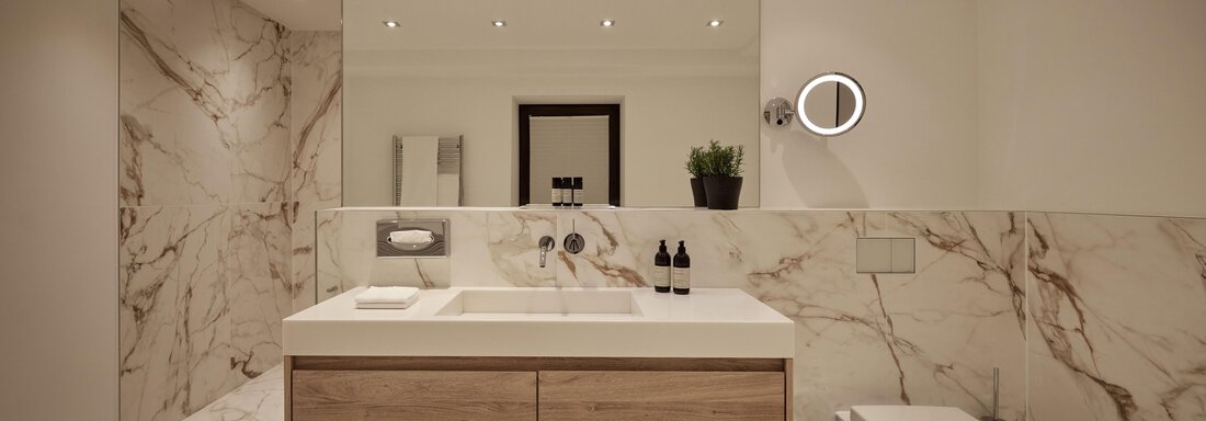 Ein modernes Badezimmer mit eleganten Marmorwänden und einem schlichten Holzwaschtisch. Im Raum befinden sich ein Spiegel, ein runder Wandspiegel und einige Pflegeprodukte. - TOP Hotel Hochgurgl - Hochgurgl