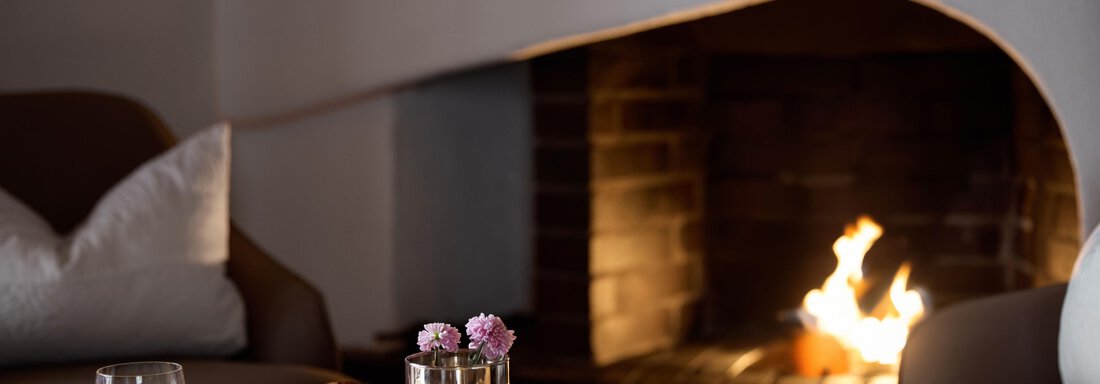 Ein gemütlicher Raum mit einem Kamin, der ein warmes Licht ausstrahlt. Auf dem Tisch stehen Getränke und Snacks, umgeben von blühenden Blumen. - TOP Hotel Hochgurgl - Hochgurgl