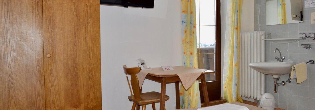 A simple guest room with two single beds, a table, and a television. The walls are light, and there is a sink as well as windows with a view. - Haus Maria - Hochsölden