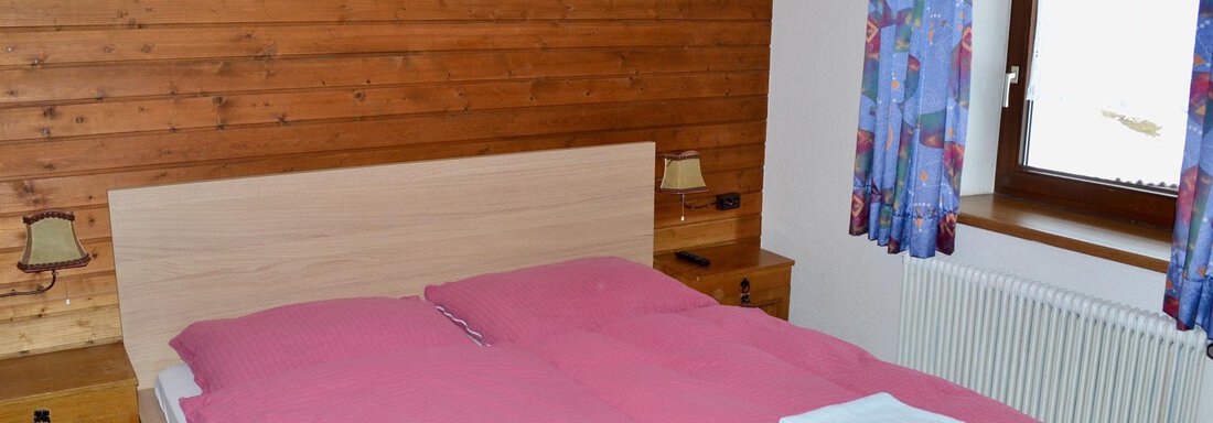 A cozy bedroom with a double bed and pink bedding. The walls are made of wood, and the window has colorful curtains. - Haus Maria - Hochsölden