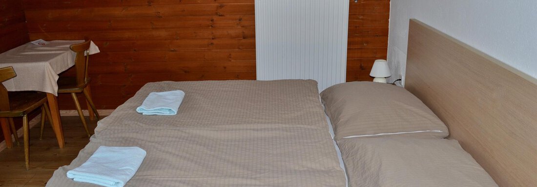 A simple bedroom with a large double bed and two pillows. In the background, a table and chairs as well as a wall heater are visible. - Haus Maria - Hochsölden