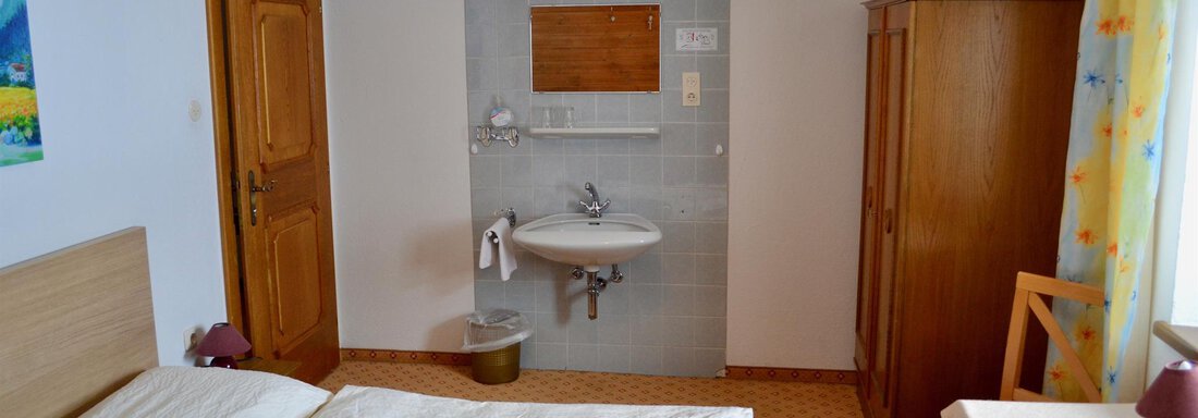 A simple room with a double bed, a table, and a chair. In the background, there is a small sink and a wooden cabinet. - Haus Maria - Hochsölden