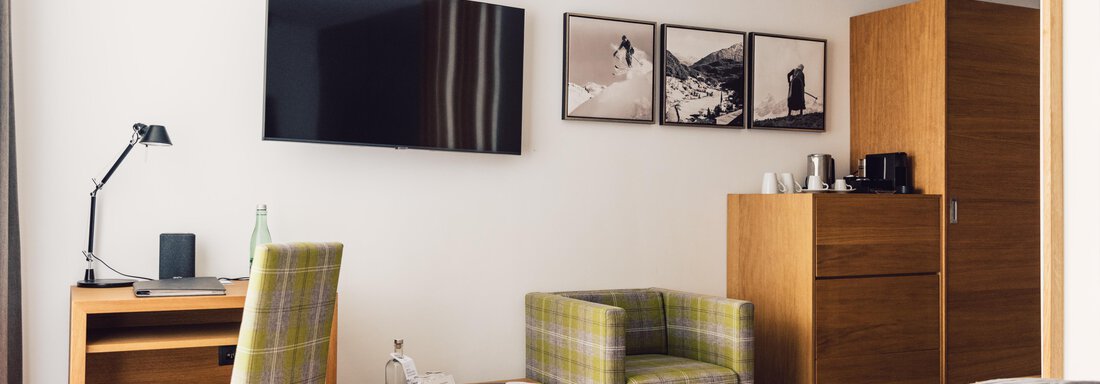 A modern hotel room with a cozy bed, a wall-mounted television, and a small desk. Two chairs and a wooden dresser complete the ambiance. - 4 * Superior Hotel Schöne Aussicht - Hochsölden