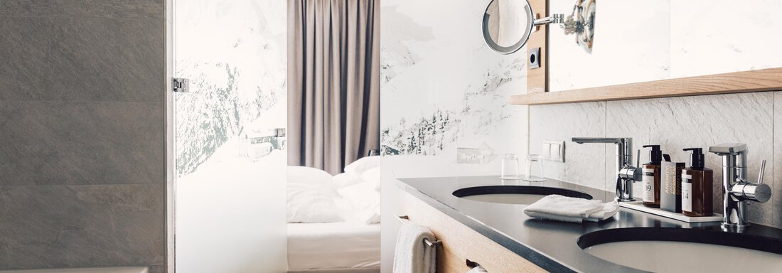 A modern bathroom with wood and marble elements. In the background, a bed and a curtain are visible. - 4 * Superior Hotel Schöne Aussicht - Hochsölden