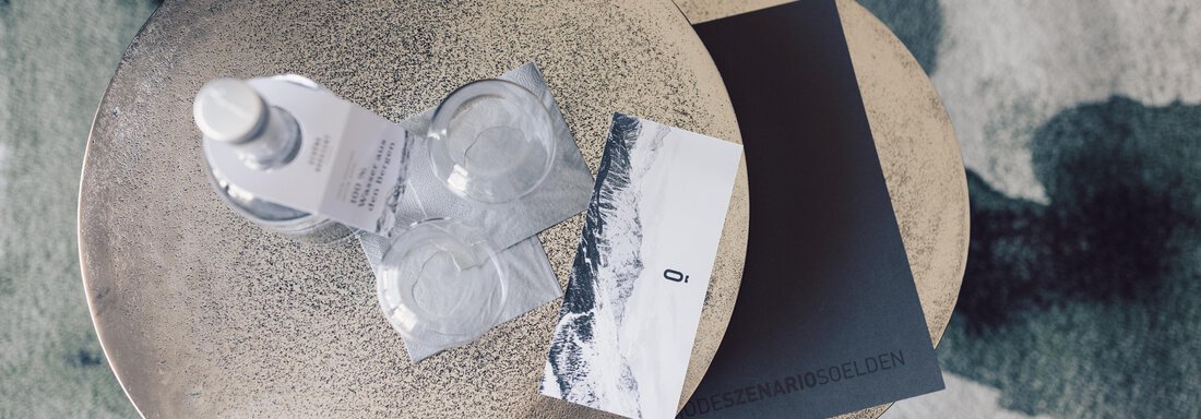 A round, bronze-colored table with a water dispenser, two glasses, and a black notebook. Next to it lies a postcard with a mountain motif. - 4 * Superior Hotel Schöne Aussicht - Hochsölden