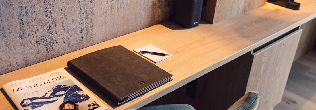 A modern desk with a wood finish. On it lies a folder, a notebook, and a pen, next to an elegant chair. - 4 * Superior Hotel Schöne Aussicht - Hochsölden
