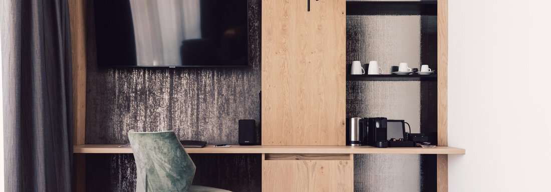 A modern room with a wooden table and a green chair. A TV hangs on the wall, and there are shelves with cups and a kettle. - 4 * Superior Hotel Schöne Aussicht - Hochsölden