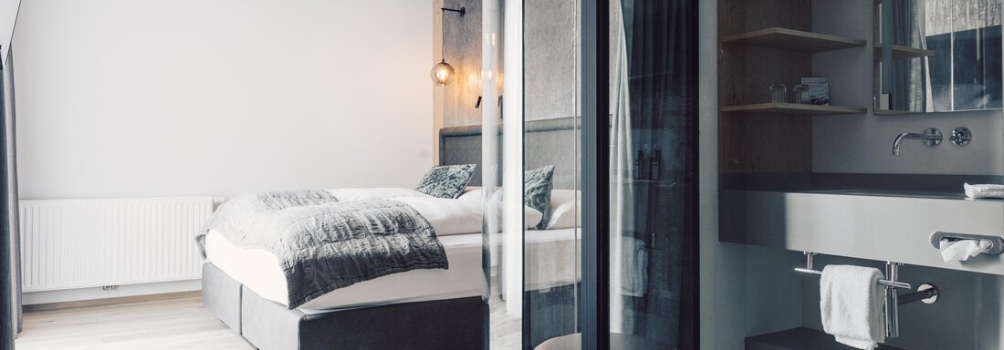 A modern bedroom with a large bed and minimalist design. In the foreground, there is an elegant bathroom with a vanity console. - 4 * Superior Hotel Schöne Aussicht - Hochsölden
