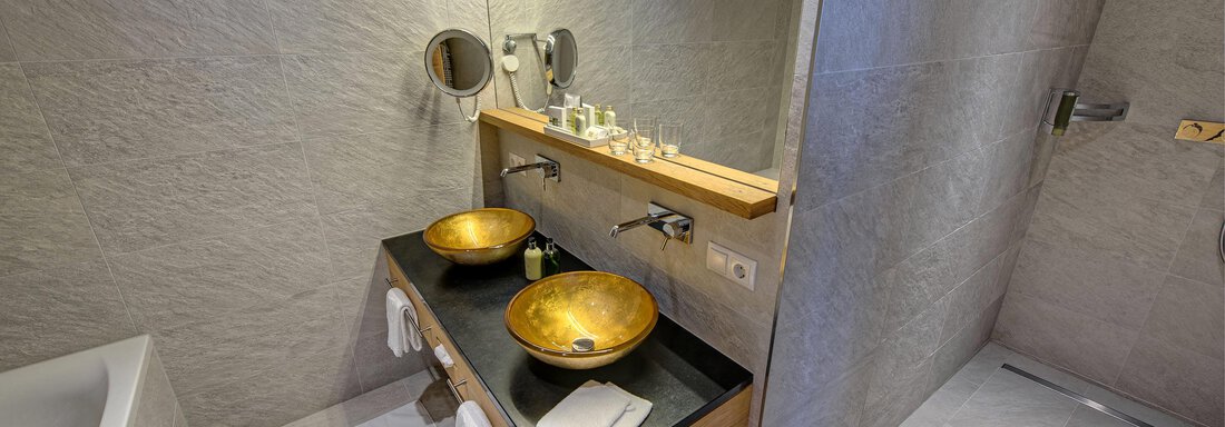 A modern bathroom with two sinks made of gold-colored metal and a wooden shelf. The walls are painted in a neutral gray, and there is plenty of light from the lighting. - 4 * Superior Hotel Schöne Aussicht - Hochsölden