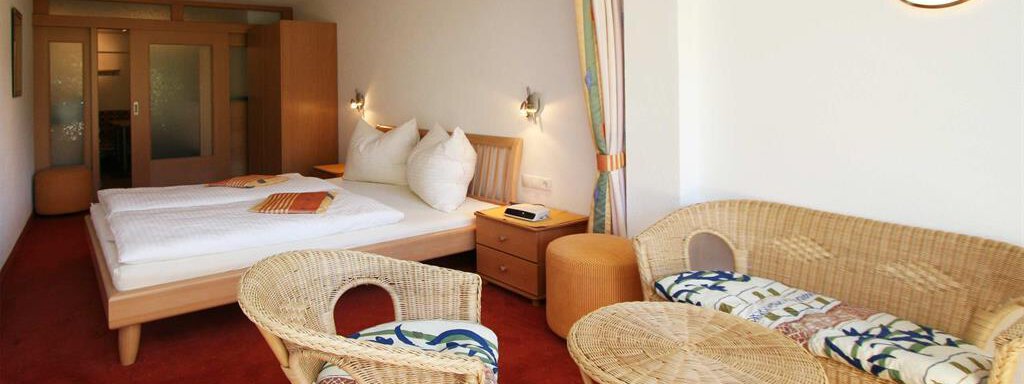 A cozy hotel room with a double bed and rattan seating. The walls are painted in light colors, and the floor is covered with red carpet. - Pension Ferdinand - Hochsölden