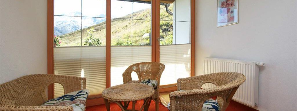 A cozy seating area with rattan furniture and a small table. Large windows provide a view of the surrounding mountains. - Pension Ferdinand - Hochsölden