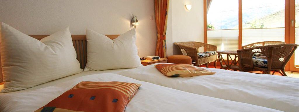 A cozy bedroom with two single beds and large pillows. Large windows provide a view of a balcony with rattan furniture. - Pension Ferdinand - Hochsölden