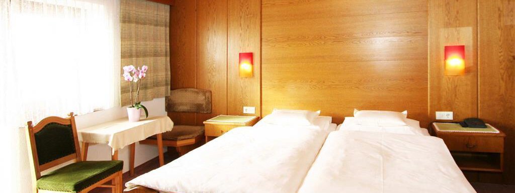 Doppelzimmer_mit_Wohnbereich A cozy bedroom with two single beds and wooden furniture. There is a table, a chair, and a large window sun that brightens the room. - Pension Ferdinand - Hochsölden
