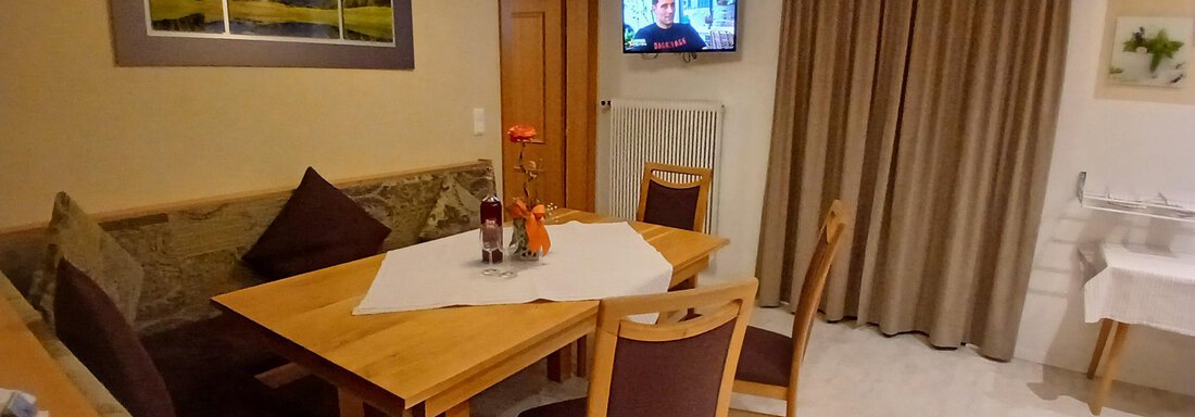 Ein gemütlicher Essbereich mit einem Tisch, Stühlen und einer Couch. Im Hintergrund ist ein Fernseher und ein Vorhang zu sehen. - Appartement Erich Wilhelm - Huben
