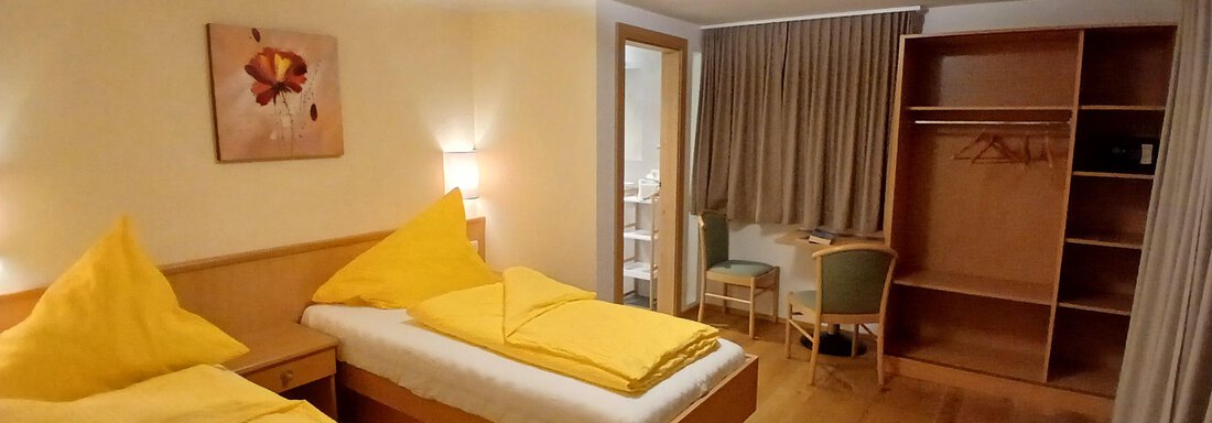 Ein helles Zimmer mit zwei Einzelbetten und gelben Kopfkissen. Es gibt einen Kleiderschrank und einen Tisch mit Stühlen. - Appartement Erich Wilhelm - Huben