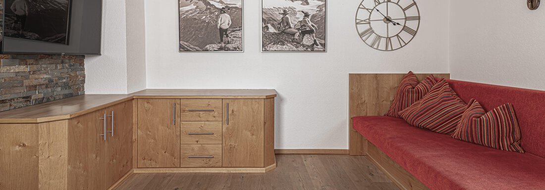 Ein gemütlicher Raum mit Holzverkleidung und einem roten Sofa. An der Wand hängen zwei Bilder und eine Uhr, die eine alpine Atmosphäre schaffen. - Appartements Alpenrose - Huben