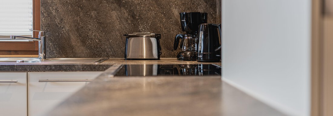 Eine moderne Küche mit einer eleganten Arbeitsplatte. Auf der Arbeitsfläche stehen ein Wasserkocher und eine Kaffeemaschine. - Appartements Alpenrose - Huben