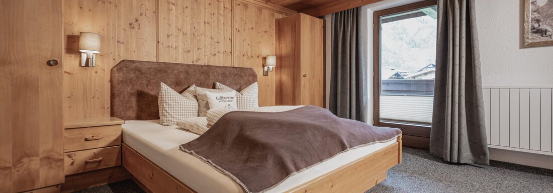 Ein gemütliches Schlafzimmer mit Holzverkleidung und einem großen Bett. Der Raum hat Fenster mit Blick nach draußen und eine warme, einladende Atmosphäre. - Appartements Alpenrose - Huben