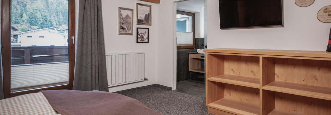 Ein gemütliches Schlafzimmer mit einem Doppelbett und einem Holzregal. Auf der anderen Seite gibt es ein Fenster mit Blick nach draußen und einen Fernseher an der Wand. - Appartements Alpenrose - Huben