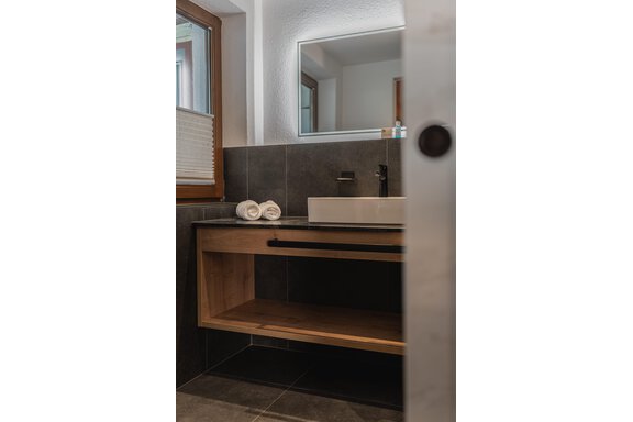 Ein modernes Badezimmer mit einem Holzwaschtisch und einer weißen Waschschüssel. Auf der Ablage liegen drei saubere Handtücher, und es gibt ein Fenster, das natürliches Licht hereinlässt. - Appartements Alpenrose - Huben