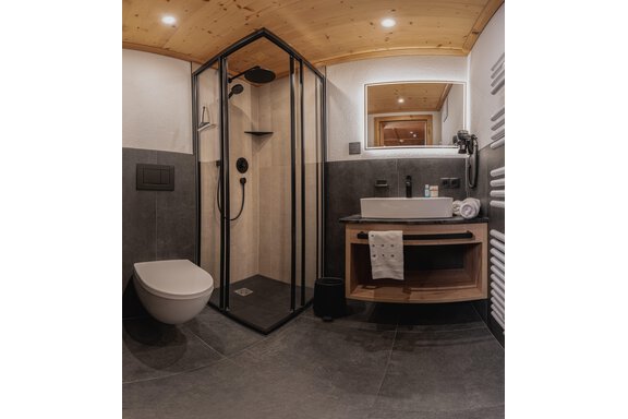 Ein modernes Badezimmer mit einer Dusche und einem modernen Waschbecken. Die Wände sind in Grautönen gehalten und es gibt eine helle Holzdecke. - Appartements Alpenrose - Huben