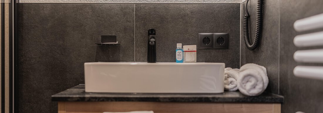 Ein modernes Badezimmer mit einem eleganten Waschbecken und Handtüchern. Die Wände sind in dunklen Farbtönen gehalten und es gibt einen großen Spiegel. - Appartements Alpenrose - Huben