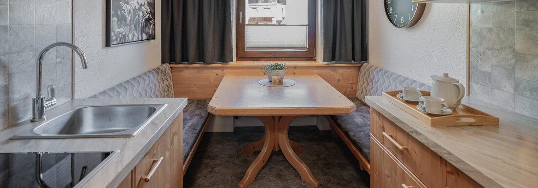 Eine gemütliche Küche mit Holzschränken und einem großen Esstisch. Die Sitzecke lädt zum Verweilen ein, und ein Fenster sorgt für Tageslicht. - Appartements Alpenrose - Huben