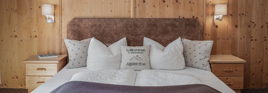 Ein gemütliches Schlafzimmer mit einem großen Bett und vielen Kissen. Die Wände sind aus Holz, und die Beleuchtung schafft eine warme Atmosphäre. - Appartements Alpenrose - Huben